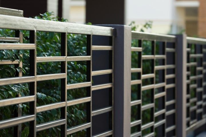 Metal fence with rectangular openings; gray and black tones. Green foliage visible behind.