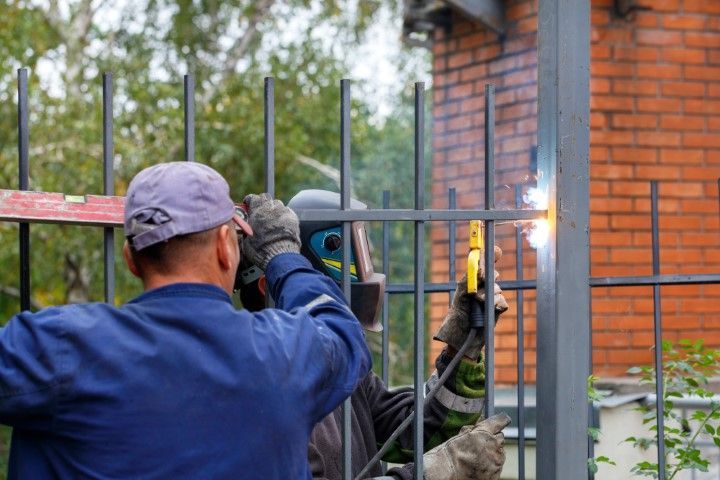 Two workers welding metal fence outdoors, one wearing a welding helmet.