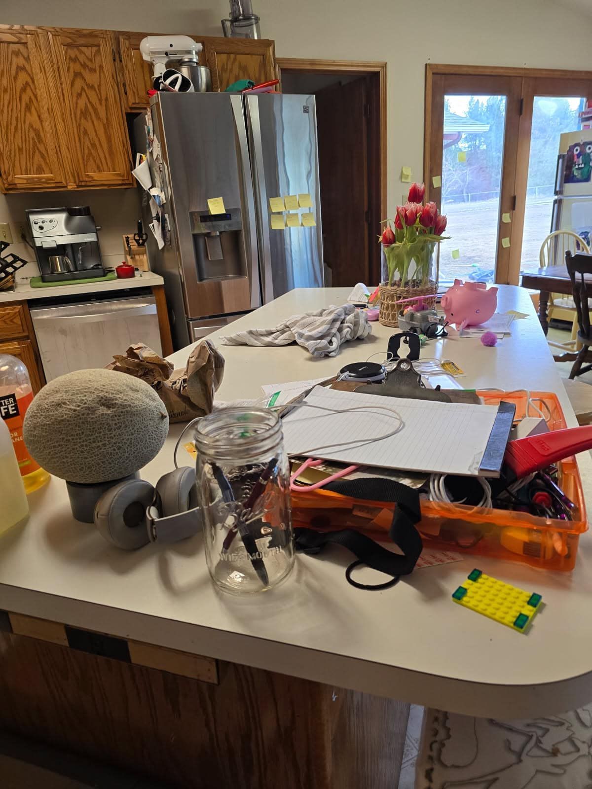 A kitchen island covered with clutter, including a jar of pens, a grey sphere, headphones, papers, and a vase of tulips.
