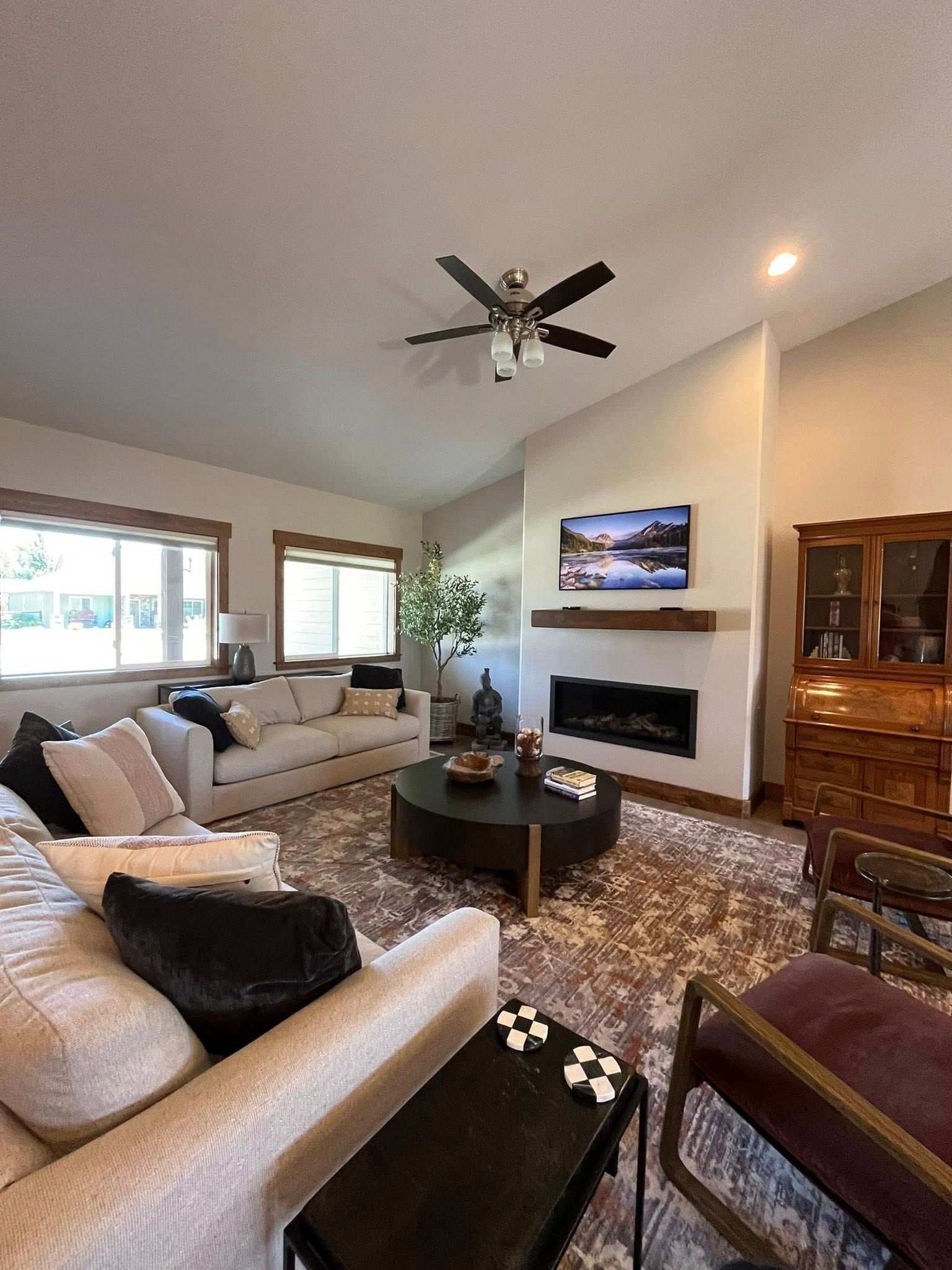 A living room with light furniture, a round black coffee table, a fireplace with a mounted TV, and large windows.