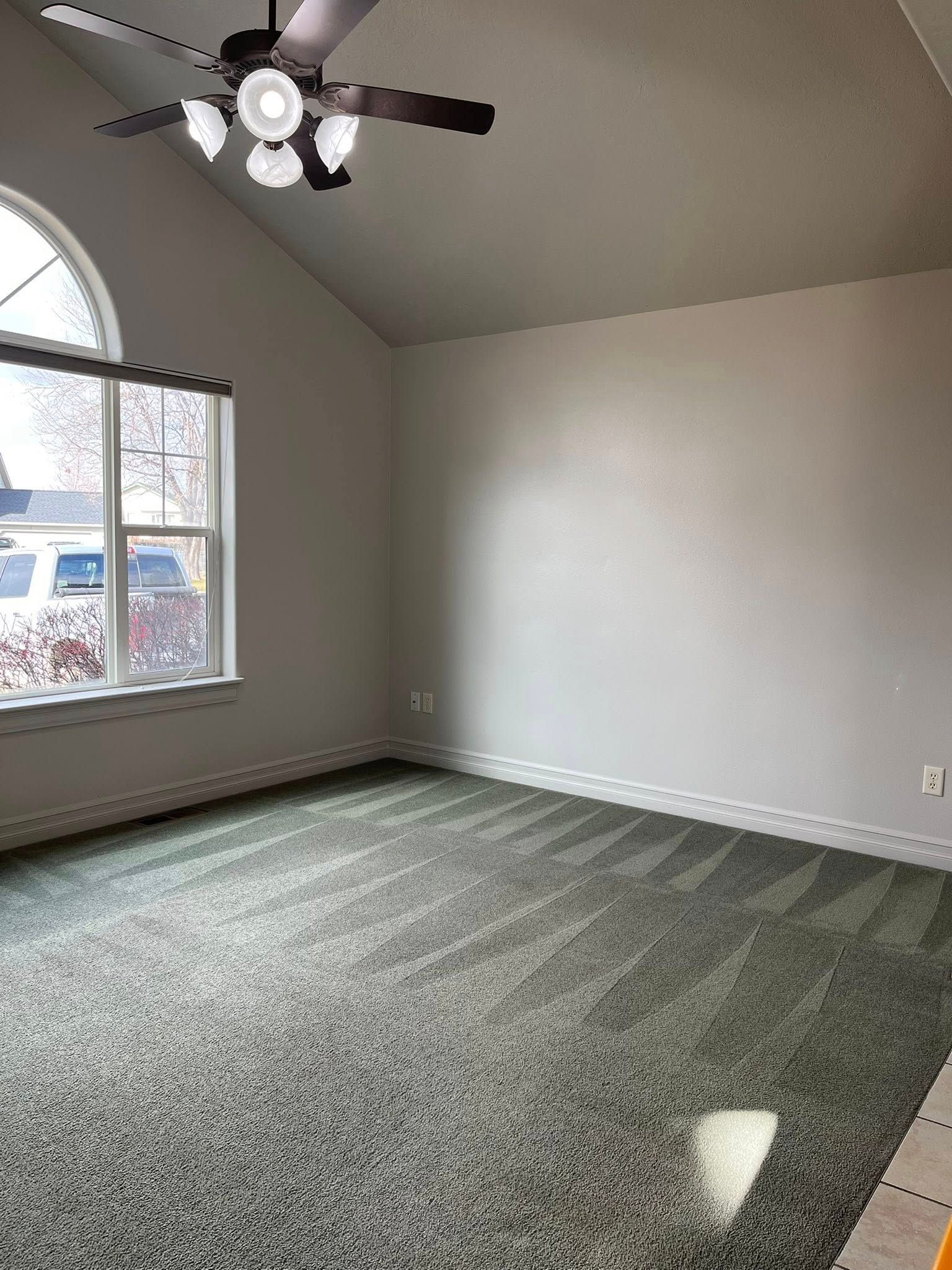 A bright, empty room featuring gray carpet, light gray walls, a vaulted ceiling, and a ceiling fan with four lights.