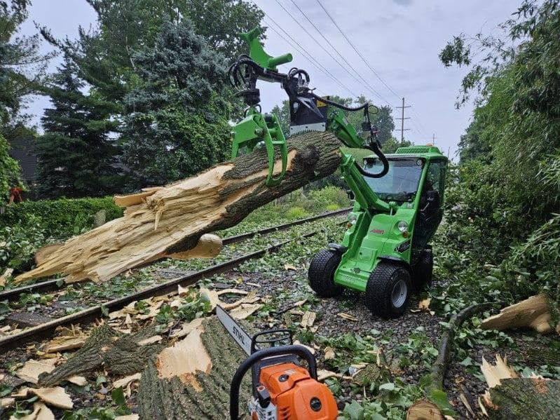Green machine with grapple arm lifting a large tree trunk near power lines and a chainsaw.
