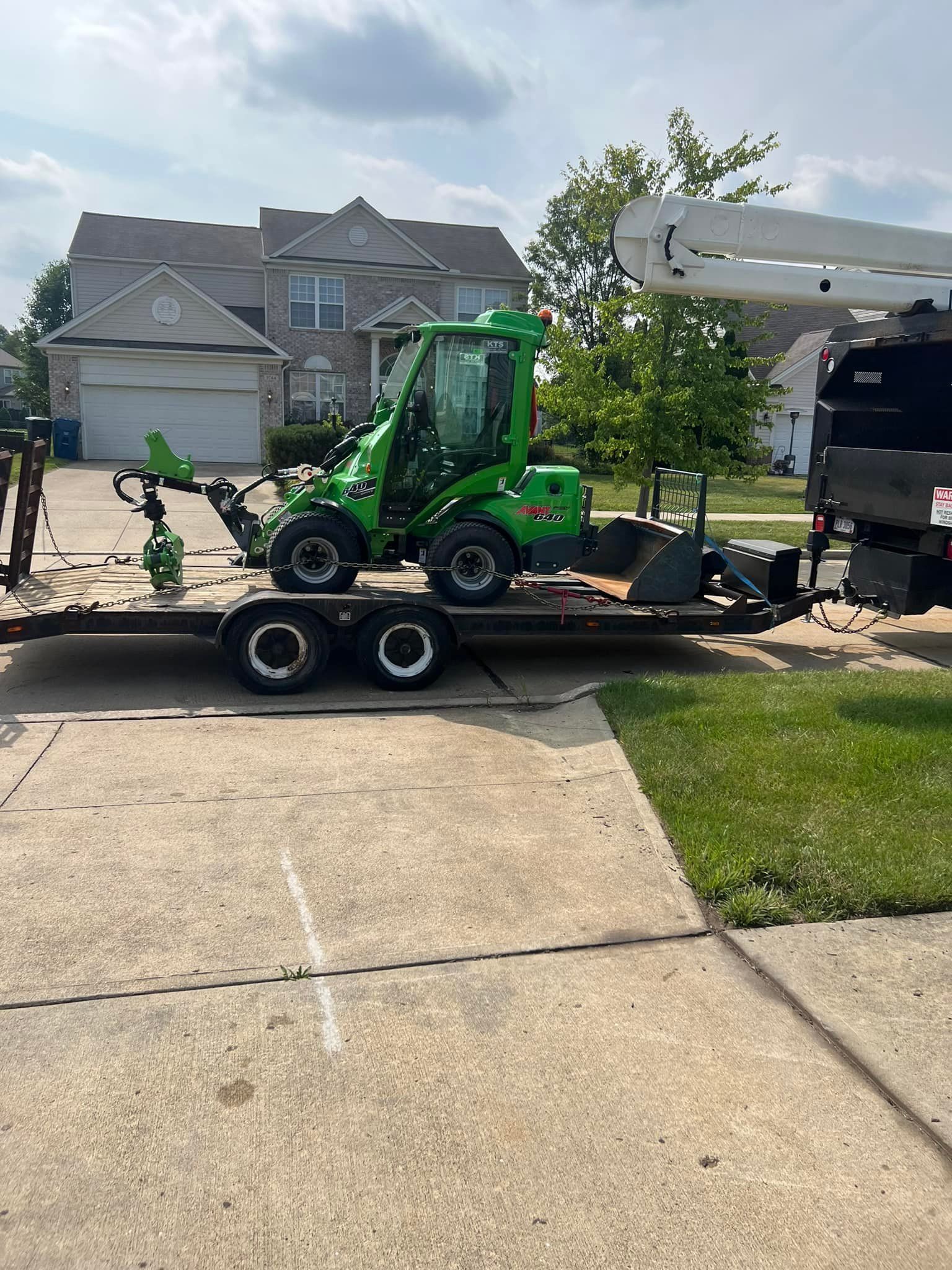 A green mini loader sits on a trailer parked in front of a two-story house on a sunny day.