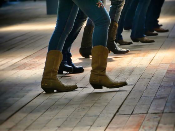 People in jeans and cowboy boots dancing on a wooden floor, with a focus on their legs and feet.