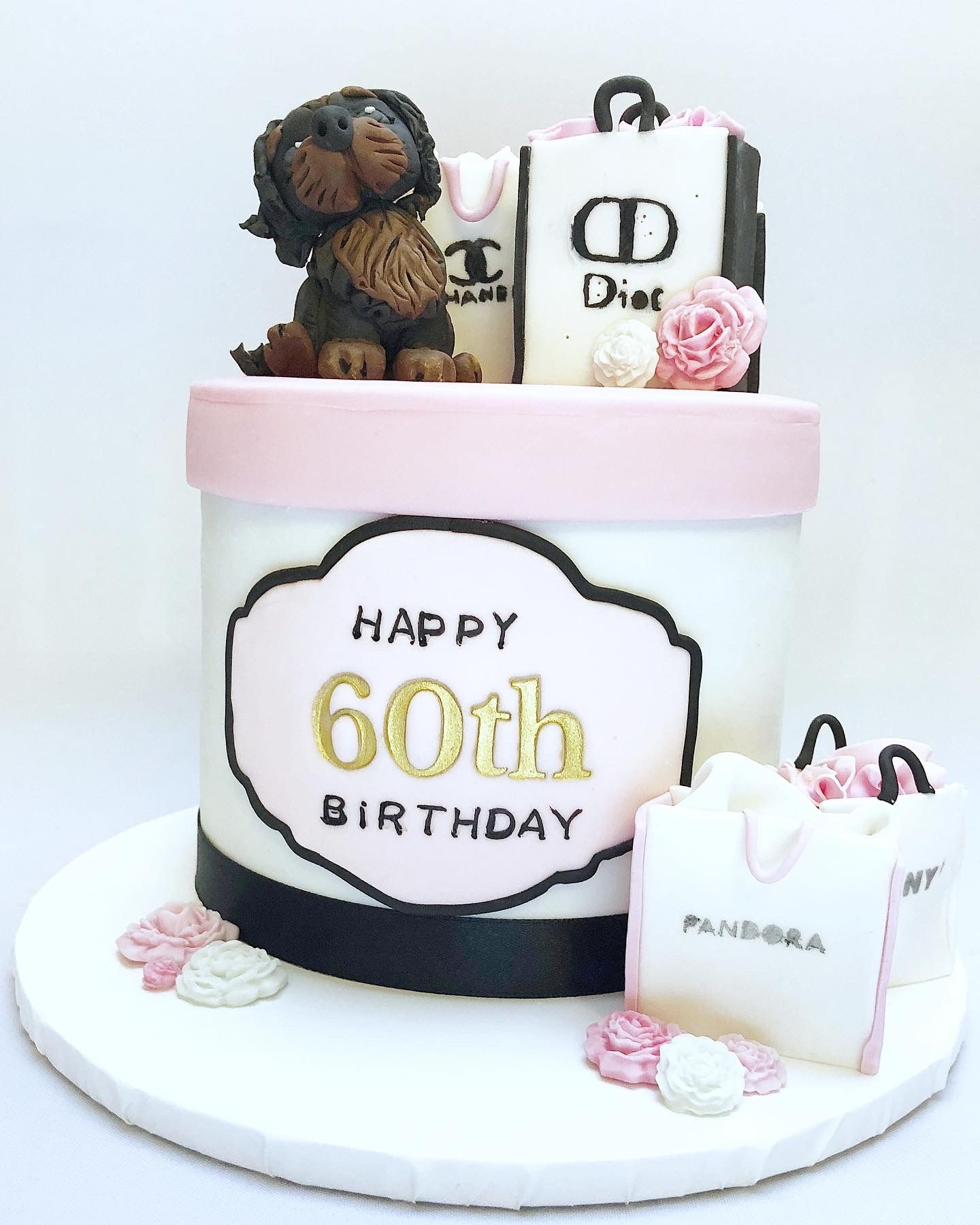 Shopaholic themed cake. The cake is beautifully decorated as a hat box in pastel pink, white and black, with Happy 60th Birthday in gold fondant on the front side of the cake. Fondant shopping bags and a little fondant dog sit atop the cake.