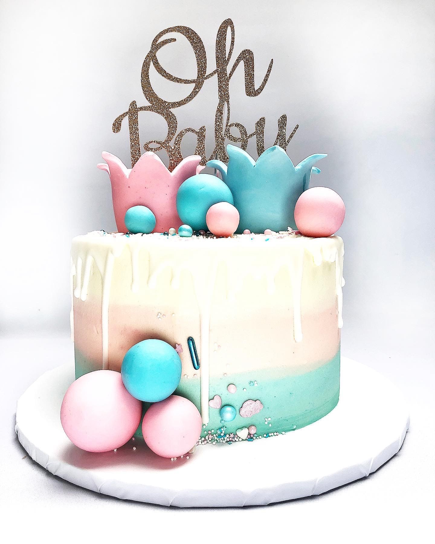 A Gender Reveal Cake with pale blue, pink and ivory ombre effect buttercream, pastel blue and pink fondant balls and a cake topper in gold reading 