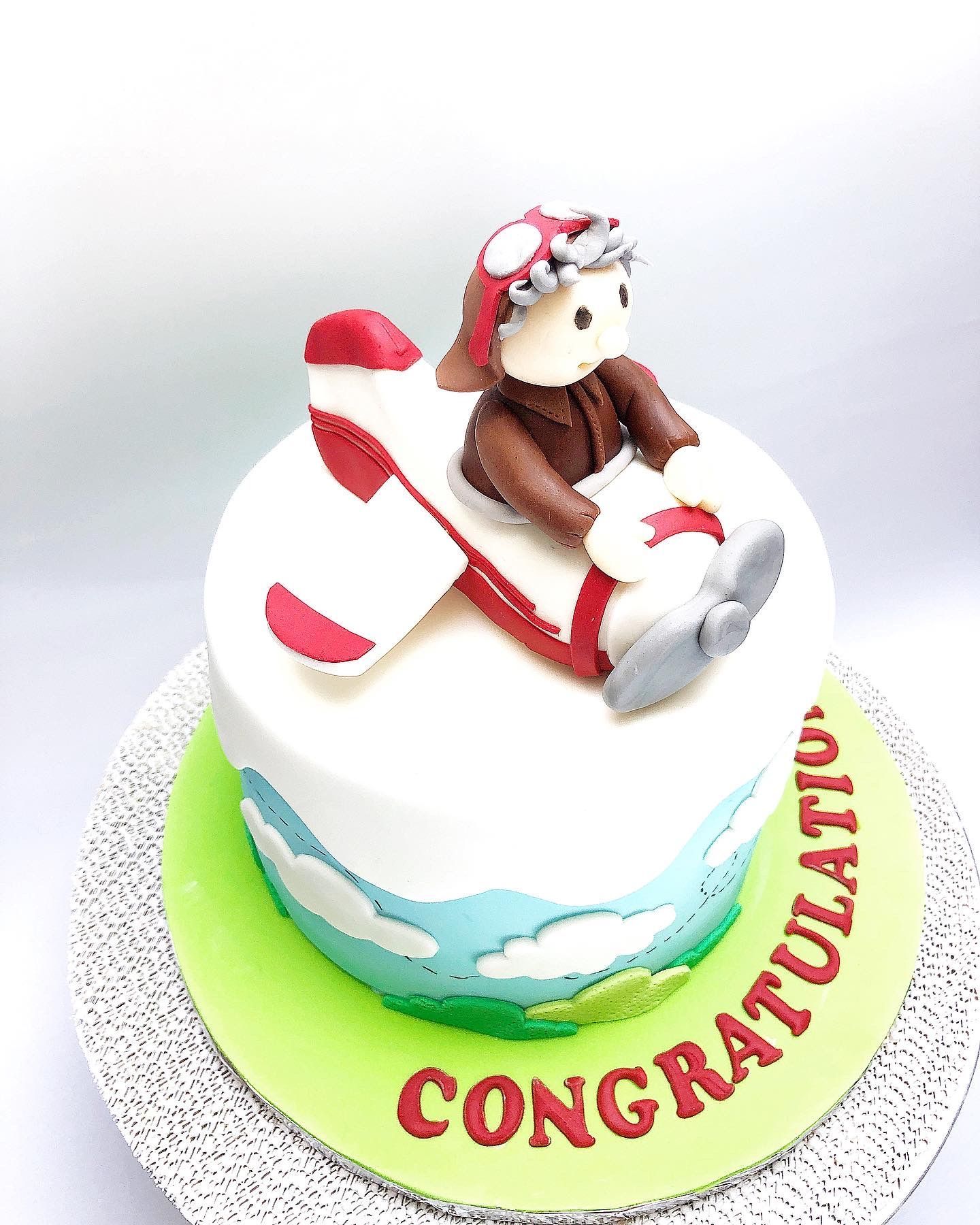 Pilot themed cake with sky, clouds and a fondant plane (with pilot) adorning the cake. The word Congratulations on the cake board.