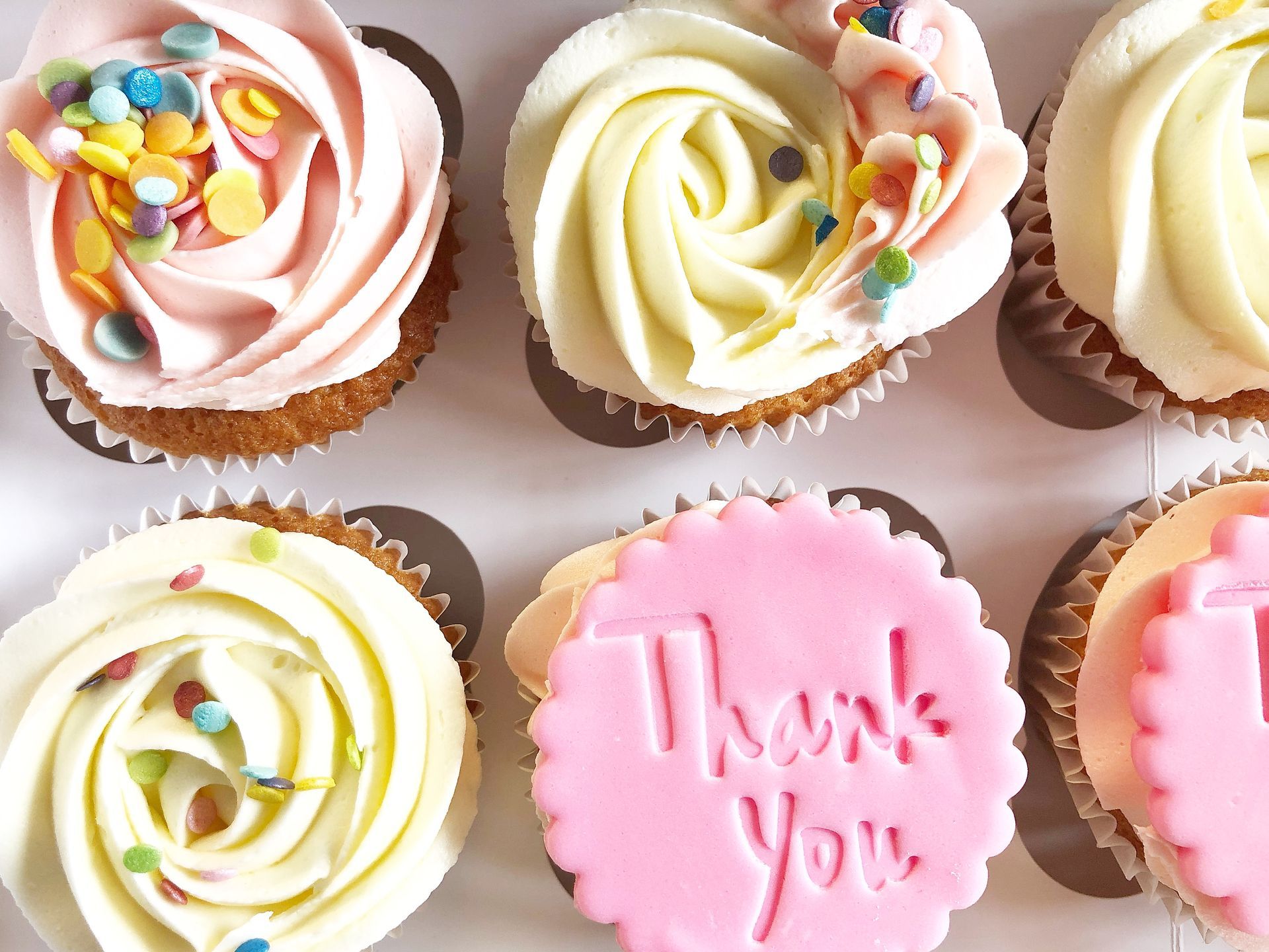 Pretty cupcakes with vanilla buttercream swirls (some pastel pink, others ivory). Confetti sprinkles and pink fondant disc saying Thank You on it.