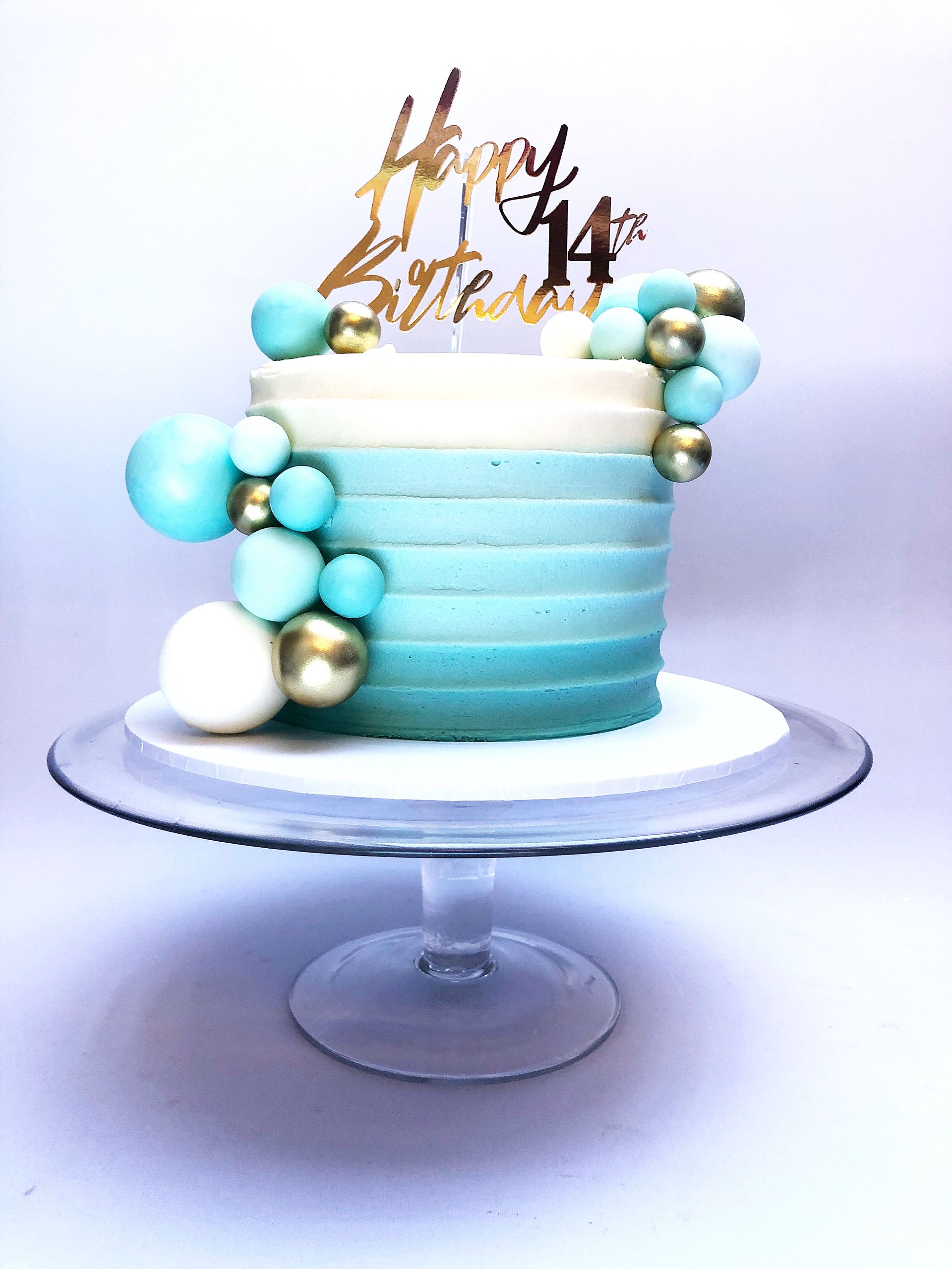 Elegant cake with blue ombre effect fading up to ivory at the top of the cake. White, gold and blue balls and a gold cake topper adorn the cake.