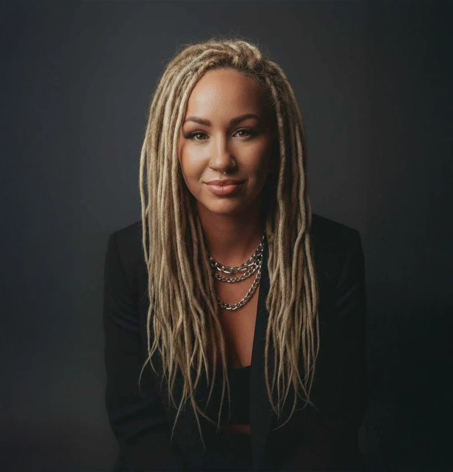 Woman with blonde dreadlocks, wearing a blazer and silver necklace, smiles at the camera against a gray backdrop.
