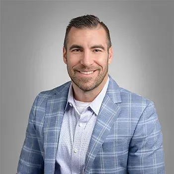 Man with a beard smiling, wearing a blue plaid jacket and button-down shirt against a gray gradient.