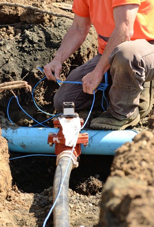 Worker connecting wires to a water valve in a trench. Orange shirt, blue pipe.