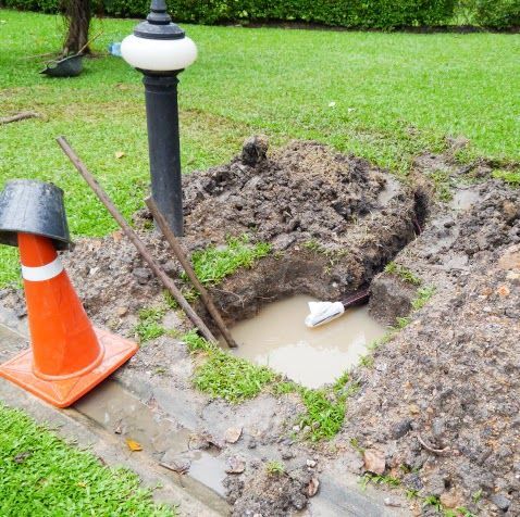 Hole dug in lawn next to street lamp, with tools, water, and orange cone nearby.