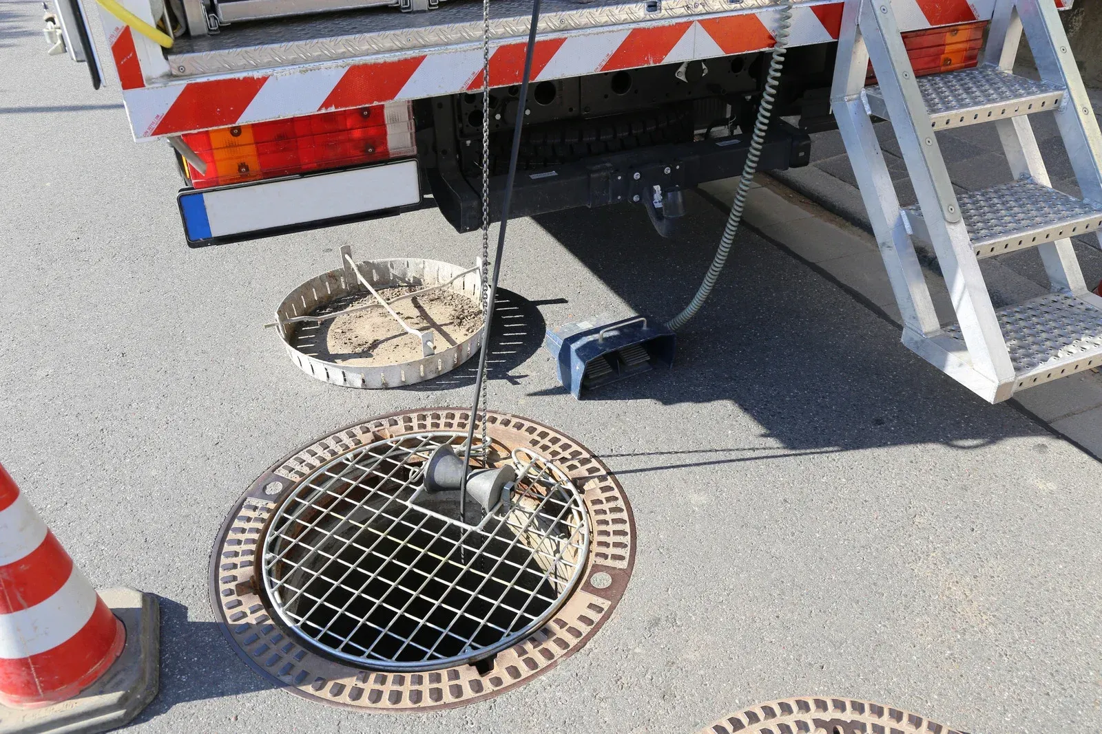 A manhole cover is open with a device suspended above it from a truck. A ladder is nearby.