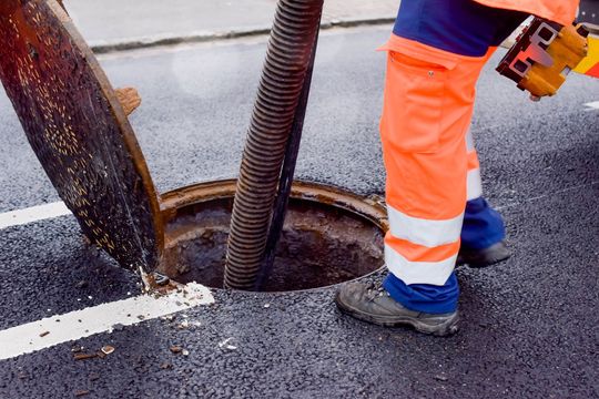 Person in orange work pants and boots, vacuuming a sewer manhole on a paved street.