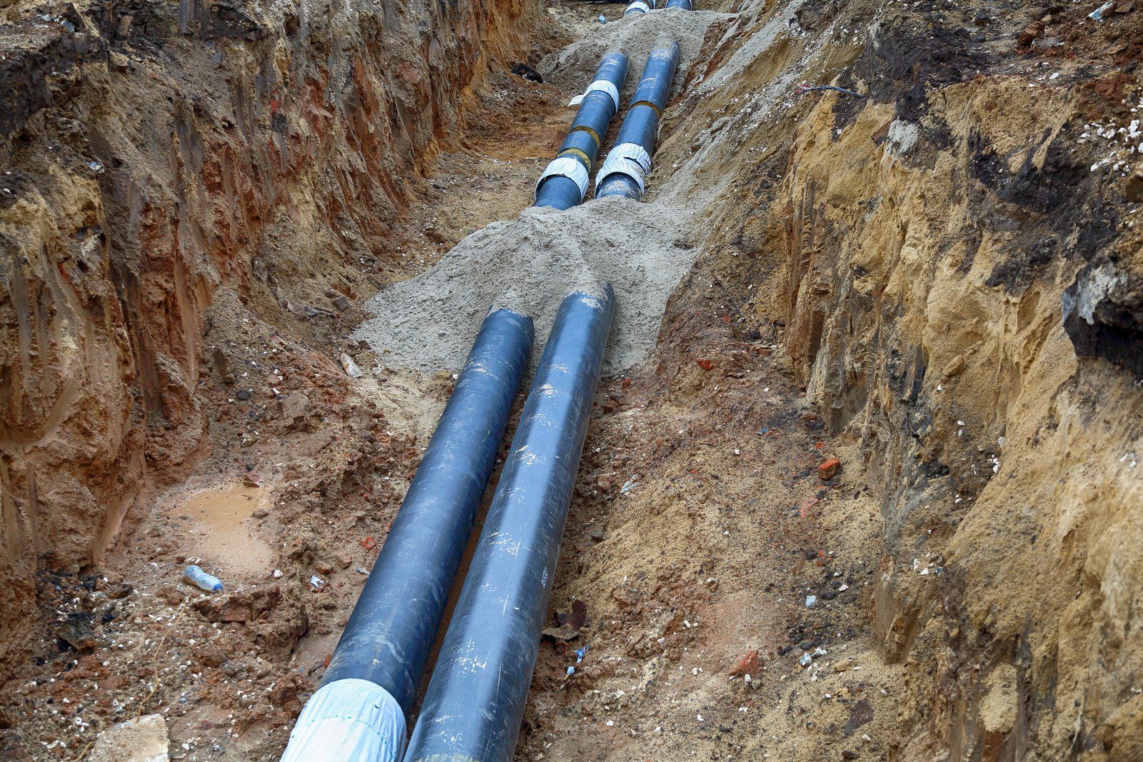 Pipes in a trench; construction site. Two parallel black pipes in the foreground, others further back. Soil surrounds.