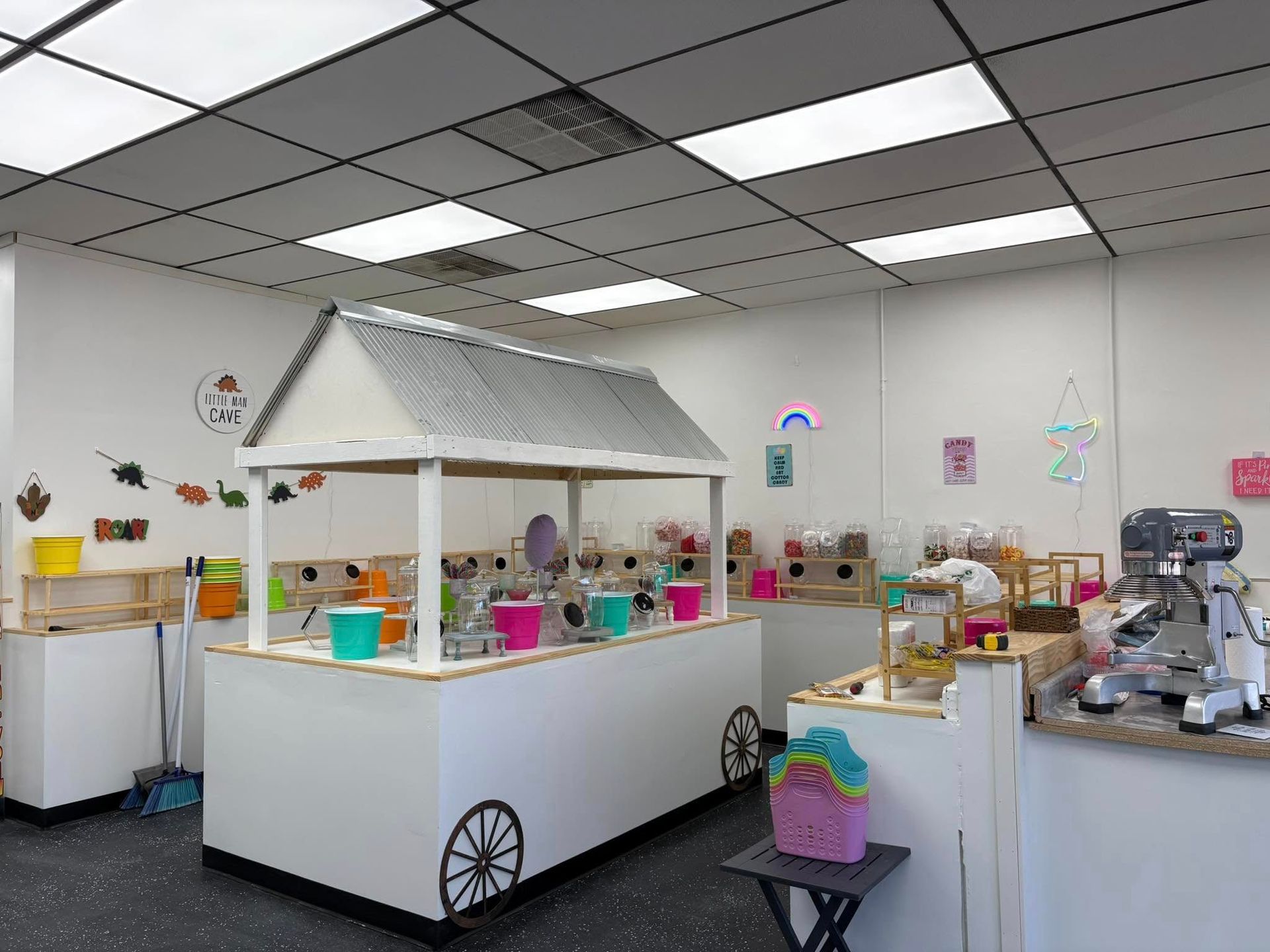 A brightly lit room features a central wooden cart with a roof, filled with colorful buckets, near a professional mixer.