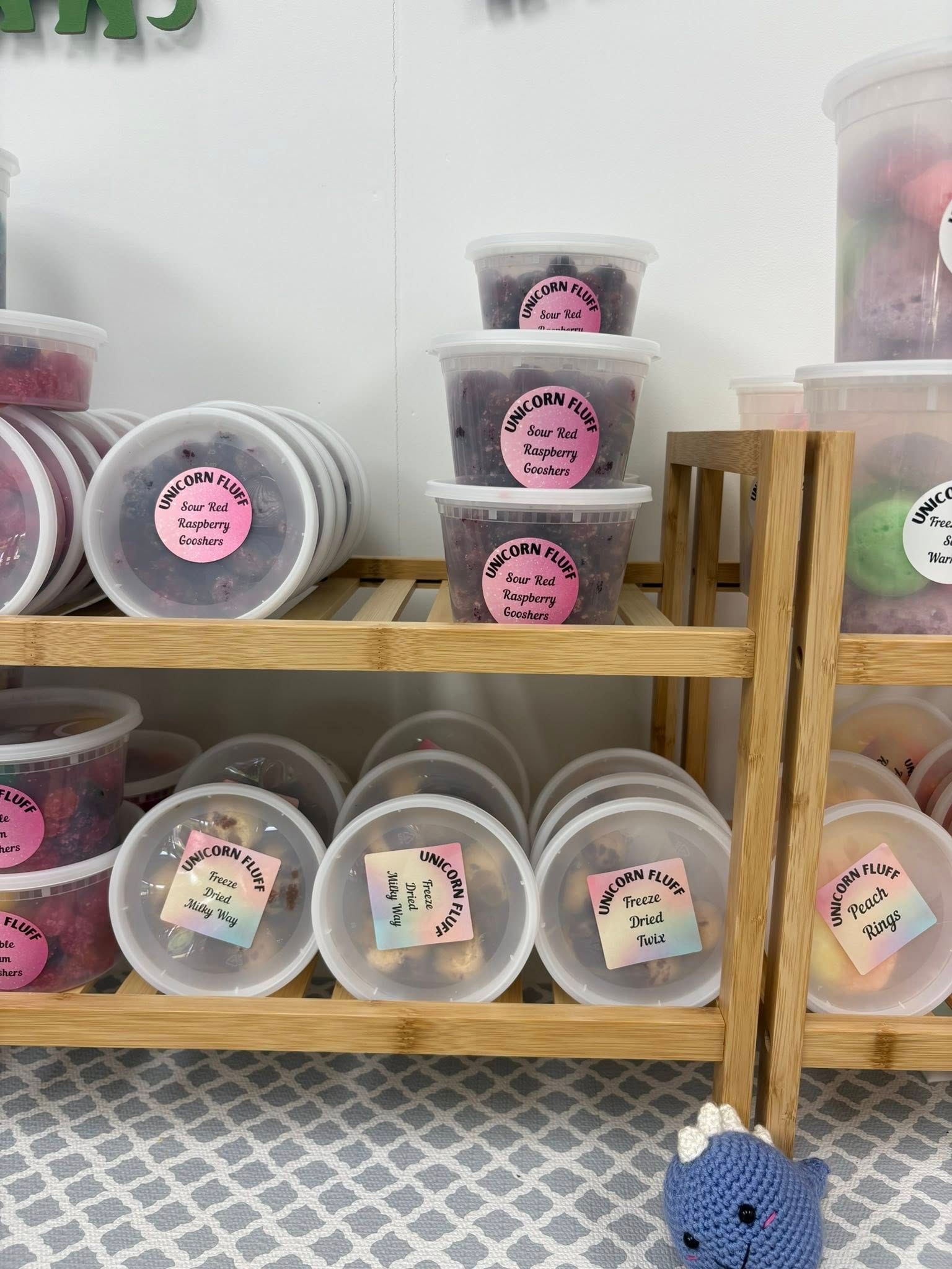 Clear plastic containers of colorful snacks labeled with stickers, displayed on a wooden shelf with a small plush toy.