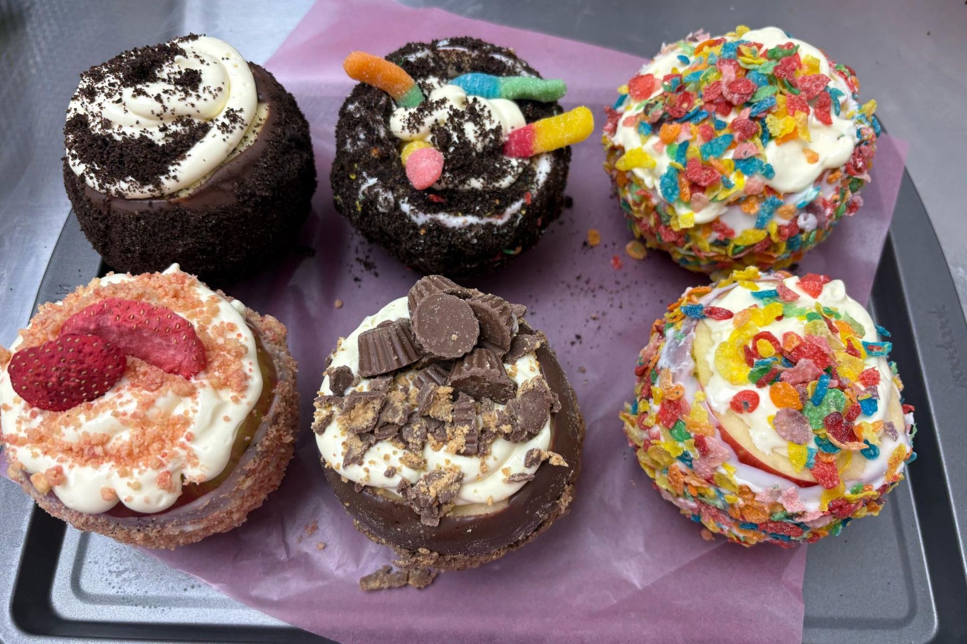 Six decorated cupcakes with various toppings like gummy worms, cereal, chocolate chunks, and strawberries on a tray.