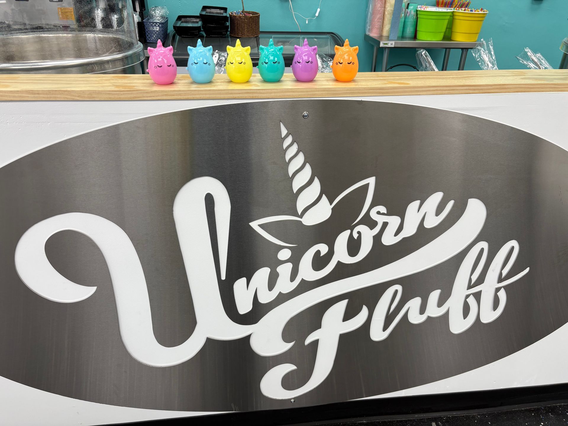 Six colorful unicorn figurines are lined up on a counter in front of a metal sign that reads