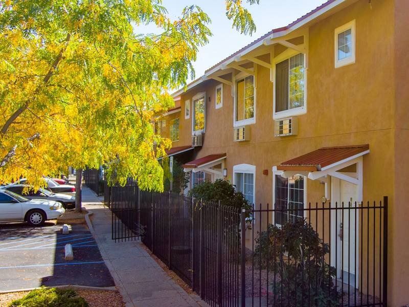 Exterior view of a brown stucco apartment building with white trim, a black metal fence, and a tree with yellow leaves at Santa Fe Suites in Santa Fe, NM.