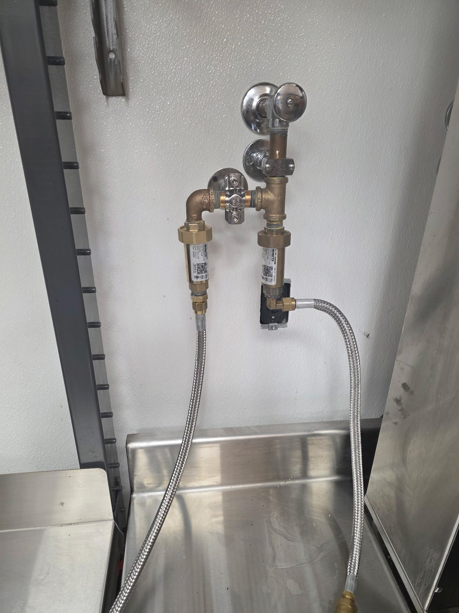 Metal plumbing fixtures with valves and braided hoses mounted above a stainless steel sink.