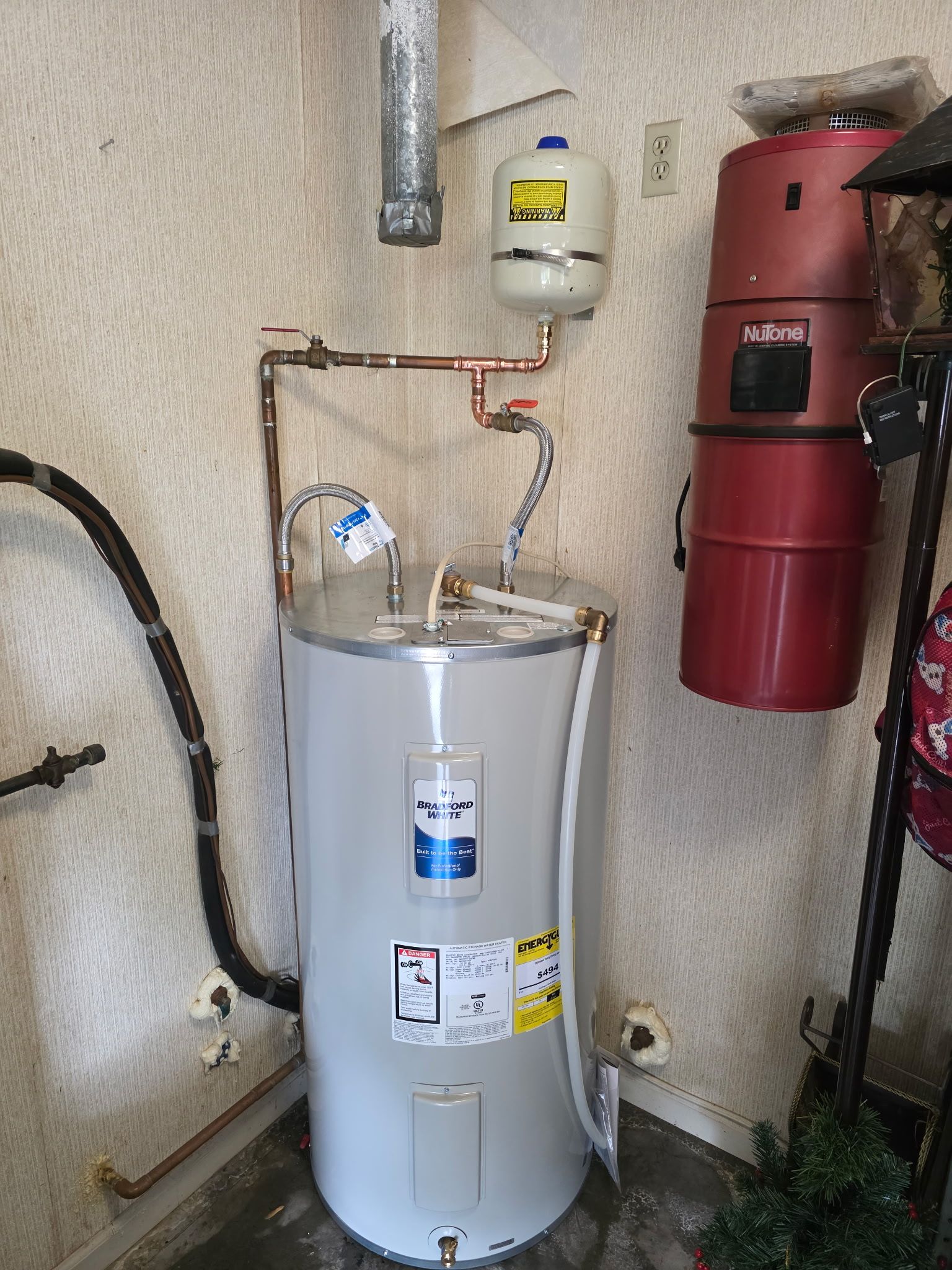 A water heater in a room with copper pipes and a red vacuum cleaner.