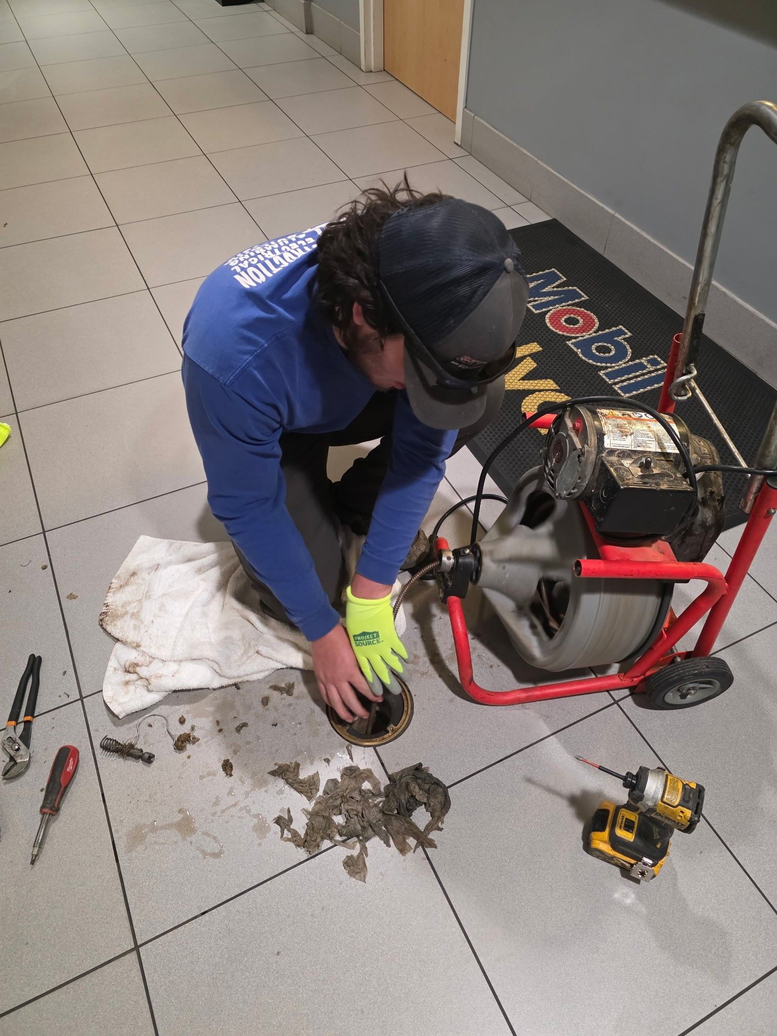 Plumber kneels on tile floor, cleaning drain with equipment; debris on floor.