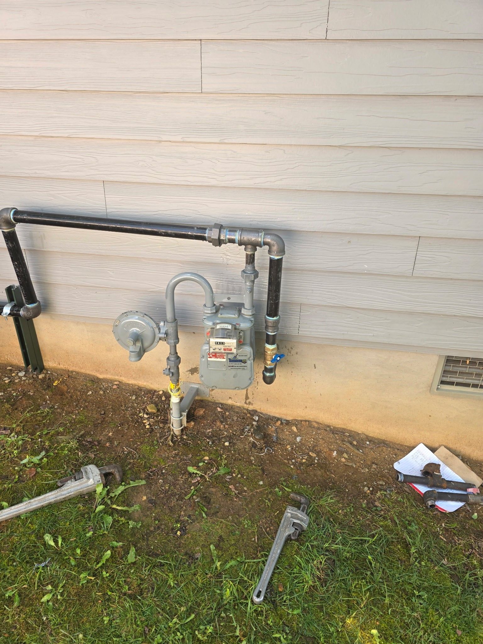 Gas meter and pipes attached to a building's exterior wall, with tools on the ground.