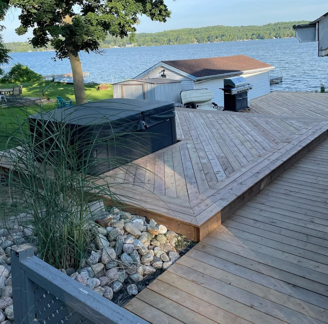 Wooden lakeside deck with lounge chair, grill, and covered boat dock beside a calm blue lake