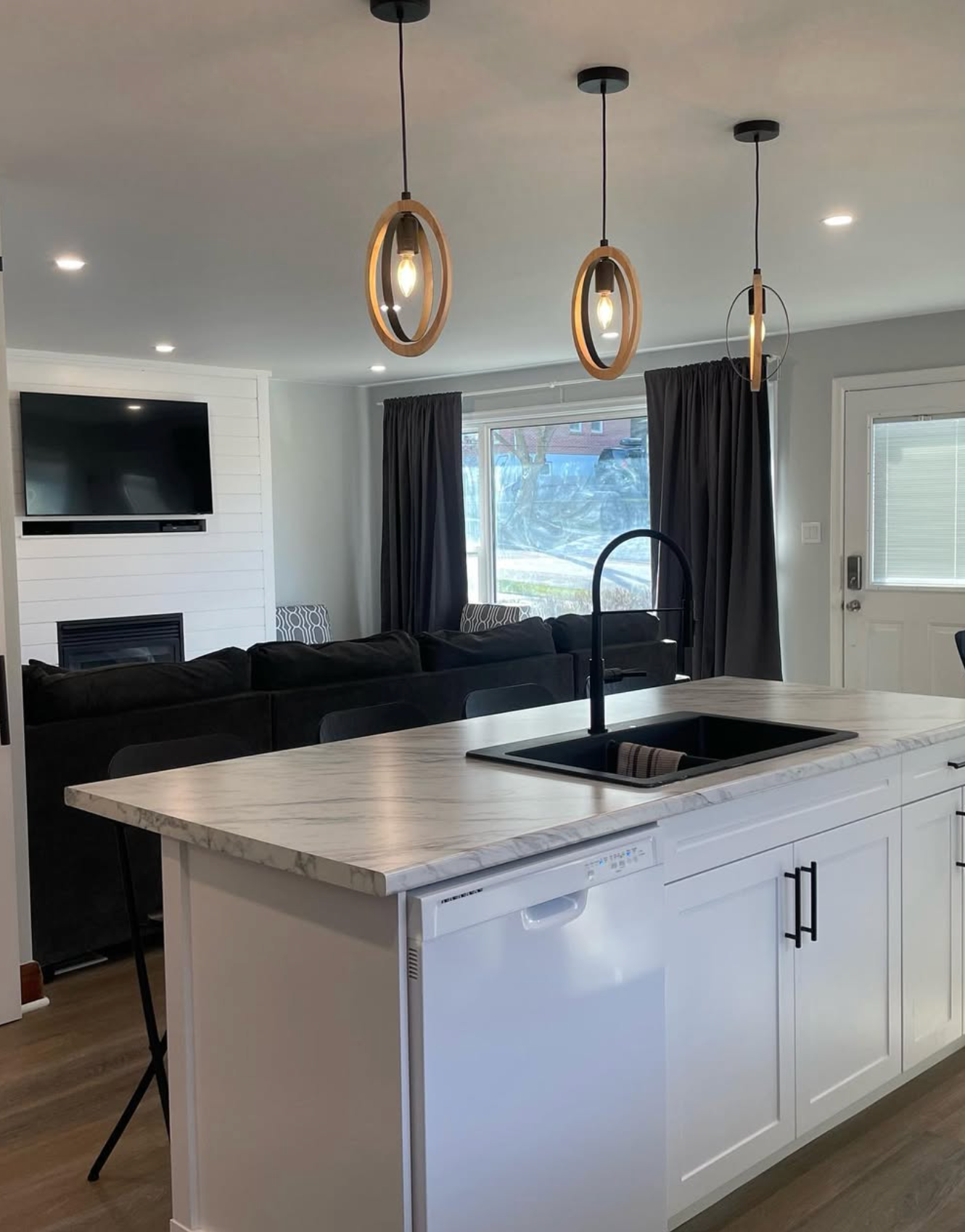 Modern kitchen with white island, black sink, and pendant lights opening to a black sofa living room