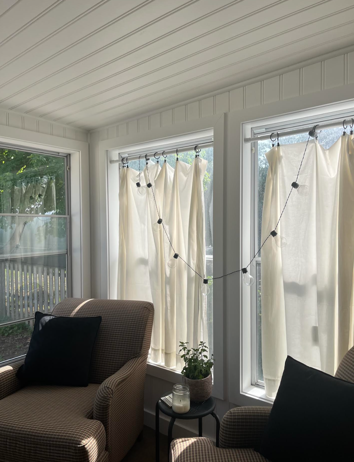 Bright sunroom with wicker chairs, white curtains, string lights, and a small table with plants and decor