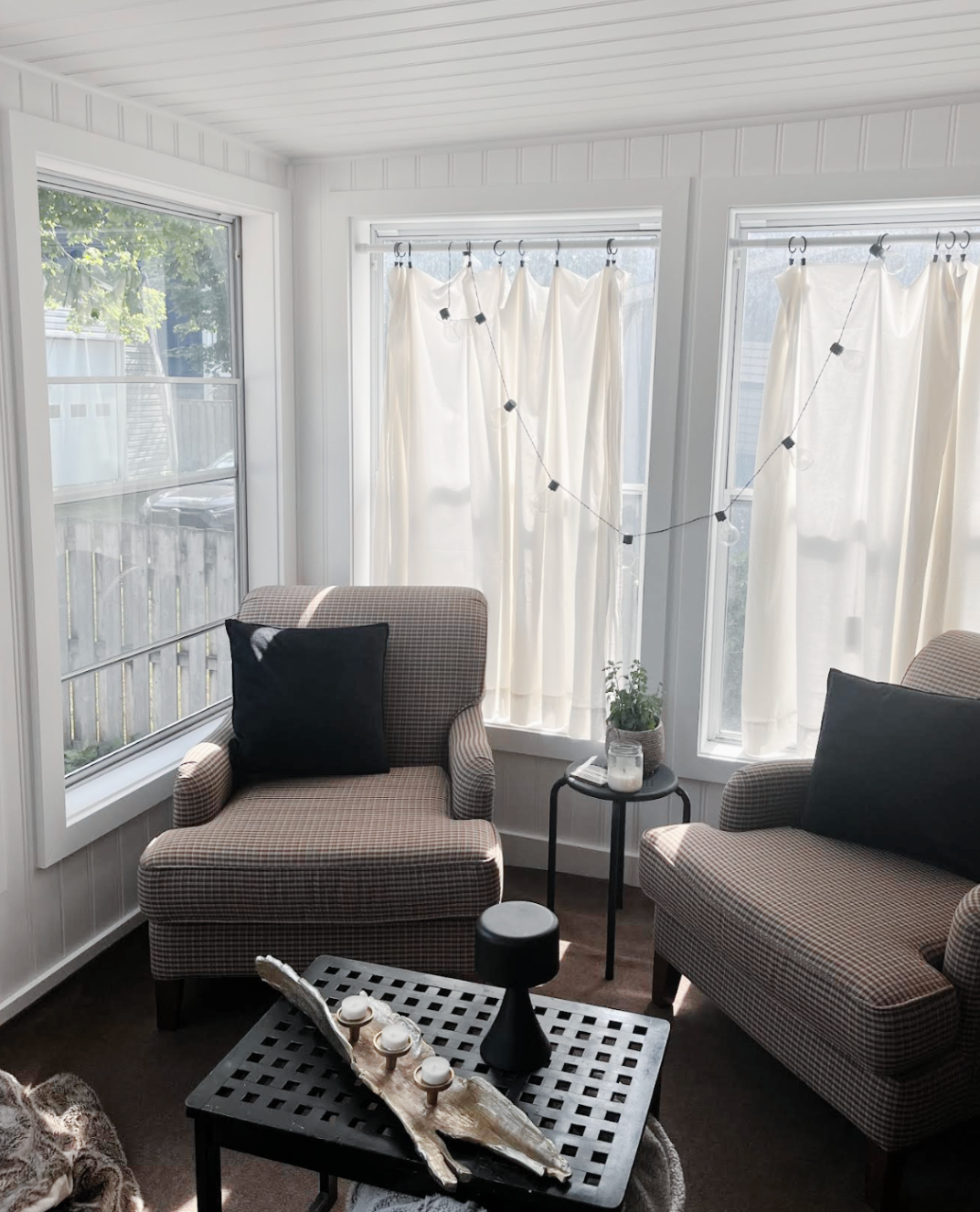 Bright sunroom with two brown chairs, black pillows, white curtains, and a small table with shells.