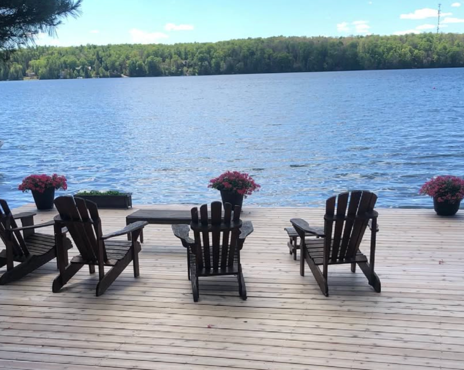 Deck chairs facing a calm blue lake, with potted flowers along a wooden dock.