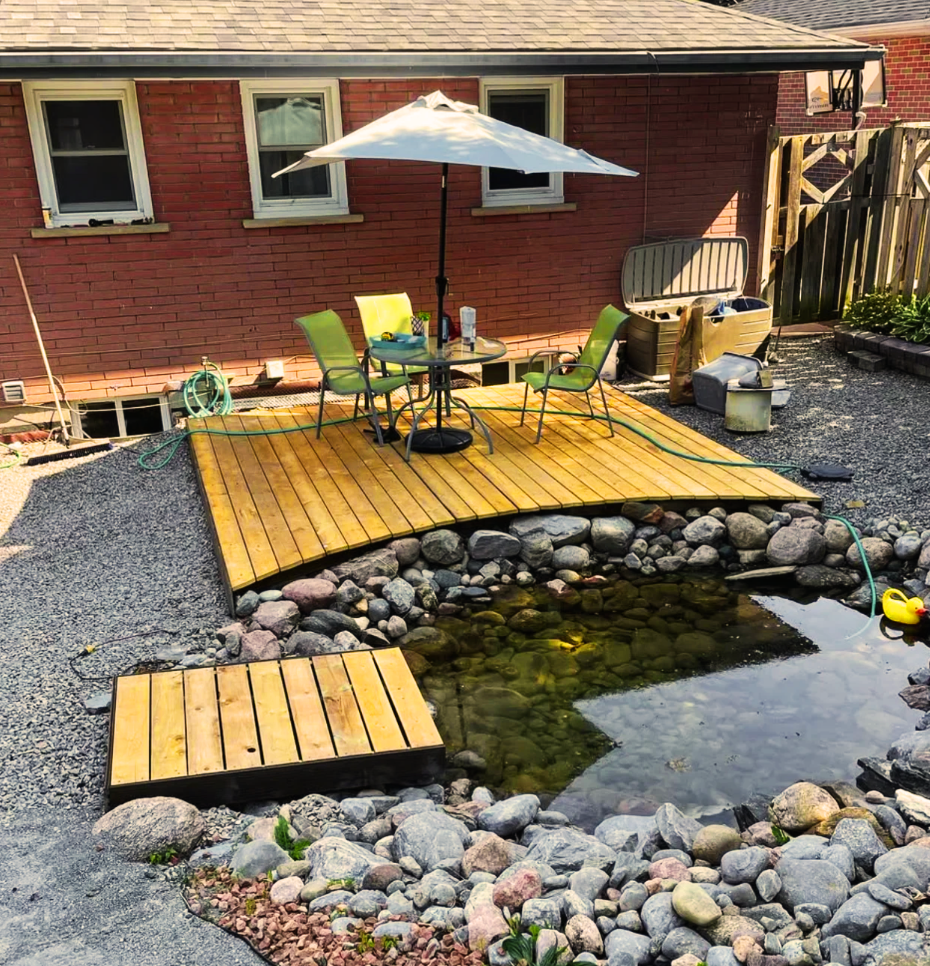 Backyard pond with wooden deck, patio table and umbrella beside a red brick house