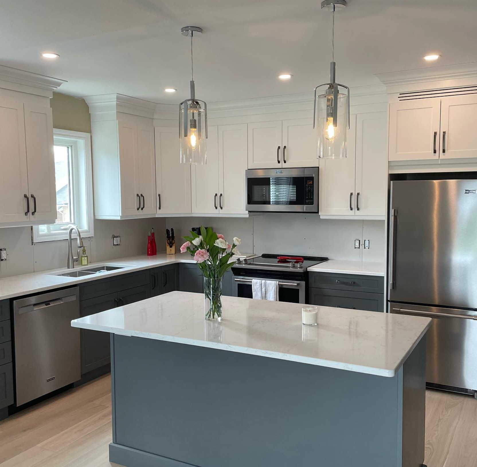 Modern white kitchen with gray island, stainless appliances, pendant lights, and a vase of flowers.
