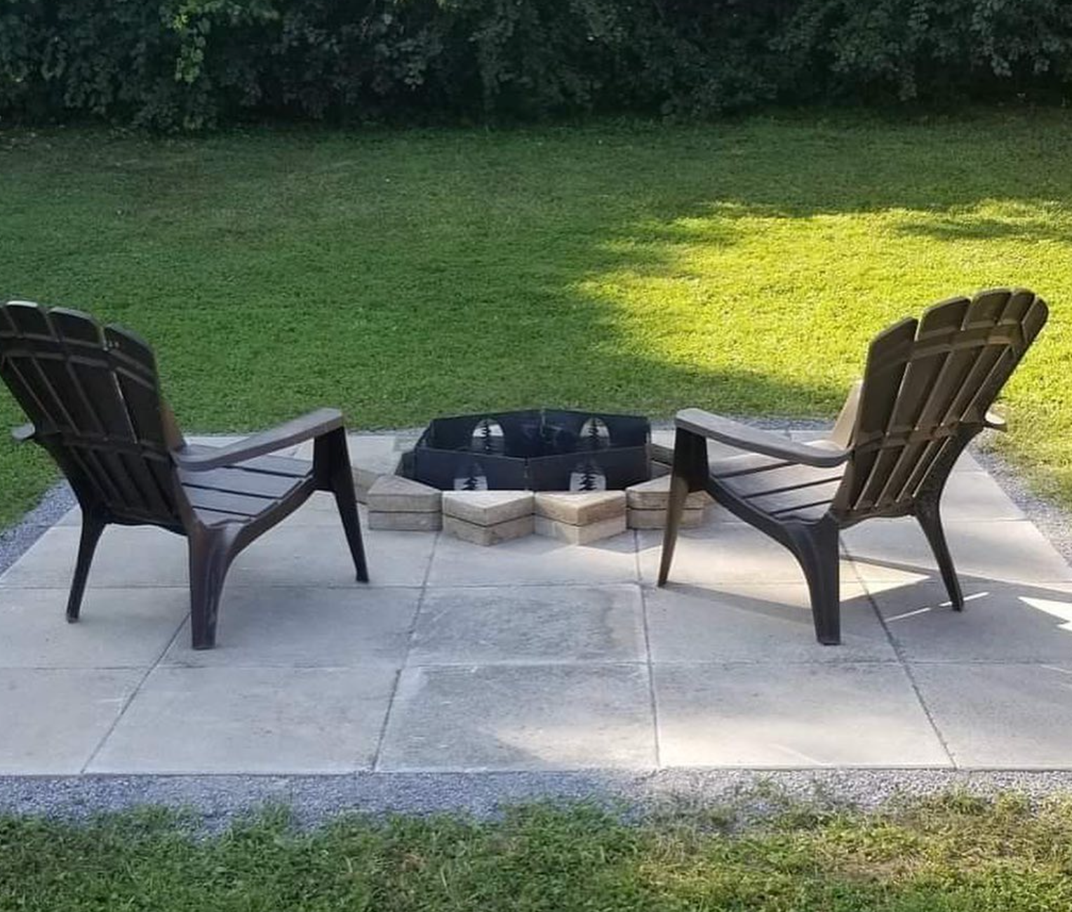 Two brown chairs facing a small fire pit on a stone patio beside a grassy lawn.
