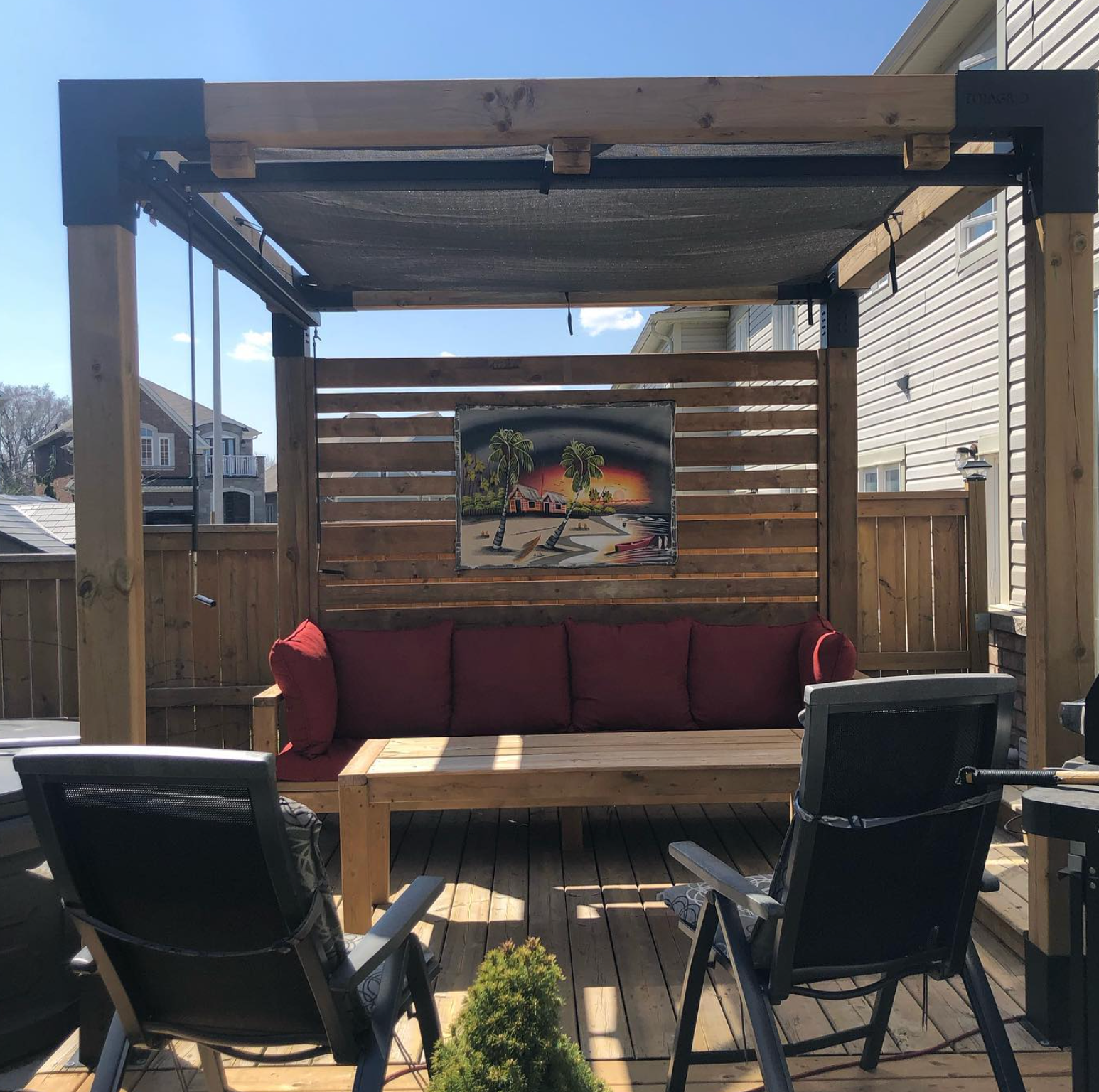 Backyard pergola with red-cushioned sofa, wooden slat wall art, and black chairs on a sunny patio