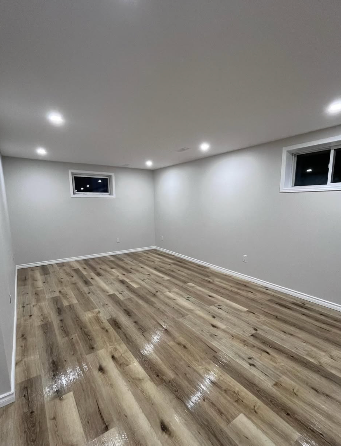 Empty finished basement room with gray walls, wood-look flooring, recessed lights, and small windows.