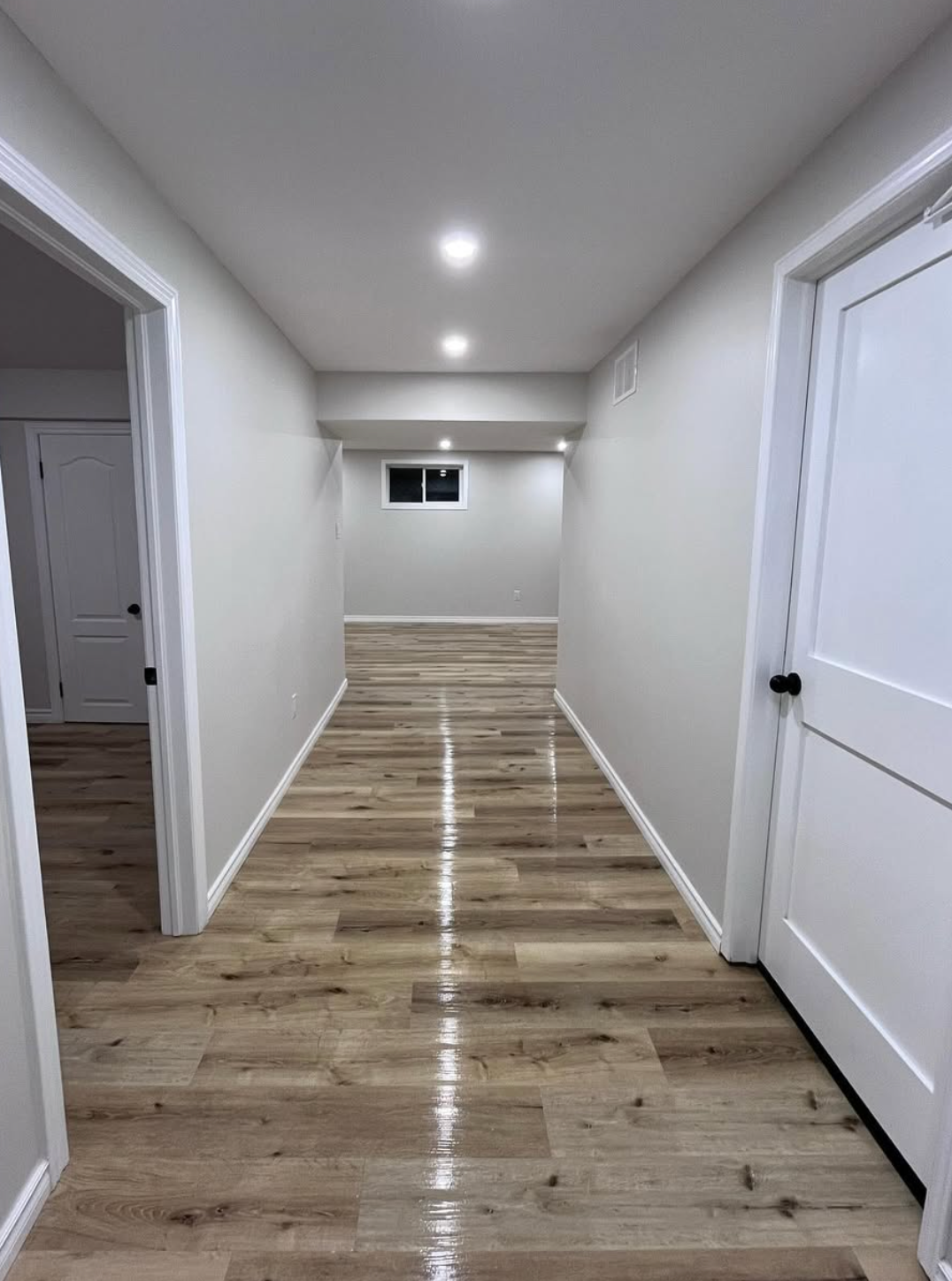 Empty hallway with white walls, wood-look floor, recessed lights, and a small window at the end.
