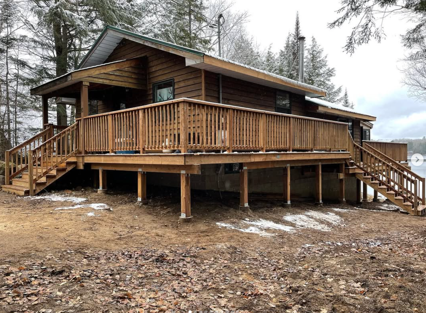 Wooden cabin with wraparound deck and stairs in a snowy forest clearing
