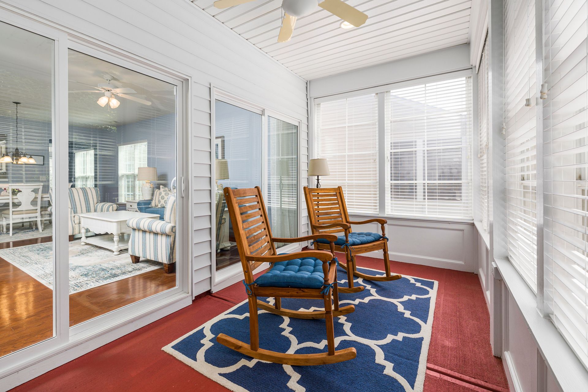 Bright sunroom with two wooden rocking chairs, blue rug, red floor, and glass doors/windows.