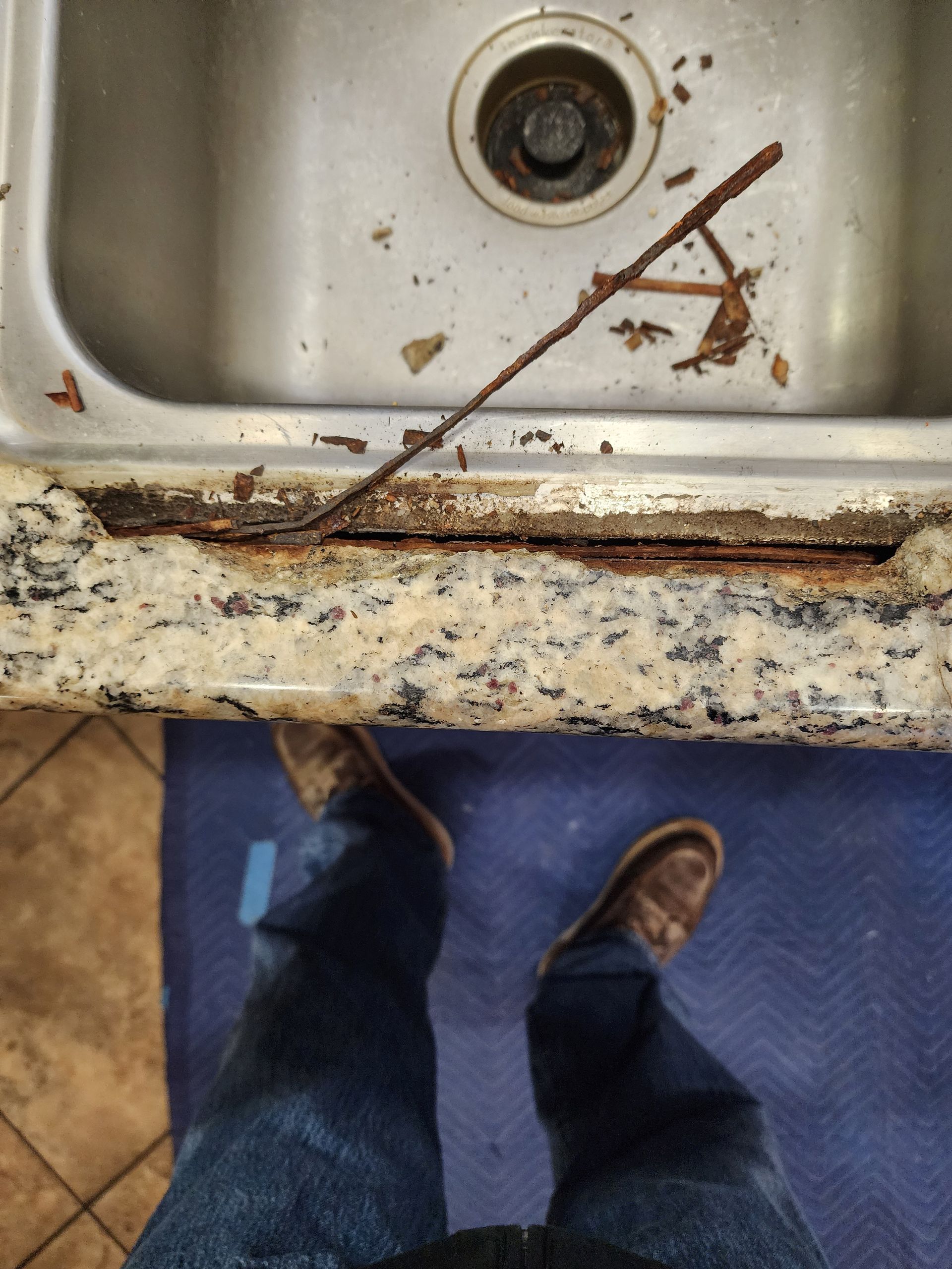 Rust-damaged countertop edge next to a stainless steel sink. Person's legs and feet are visible, wearing jeans and boots.