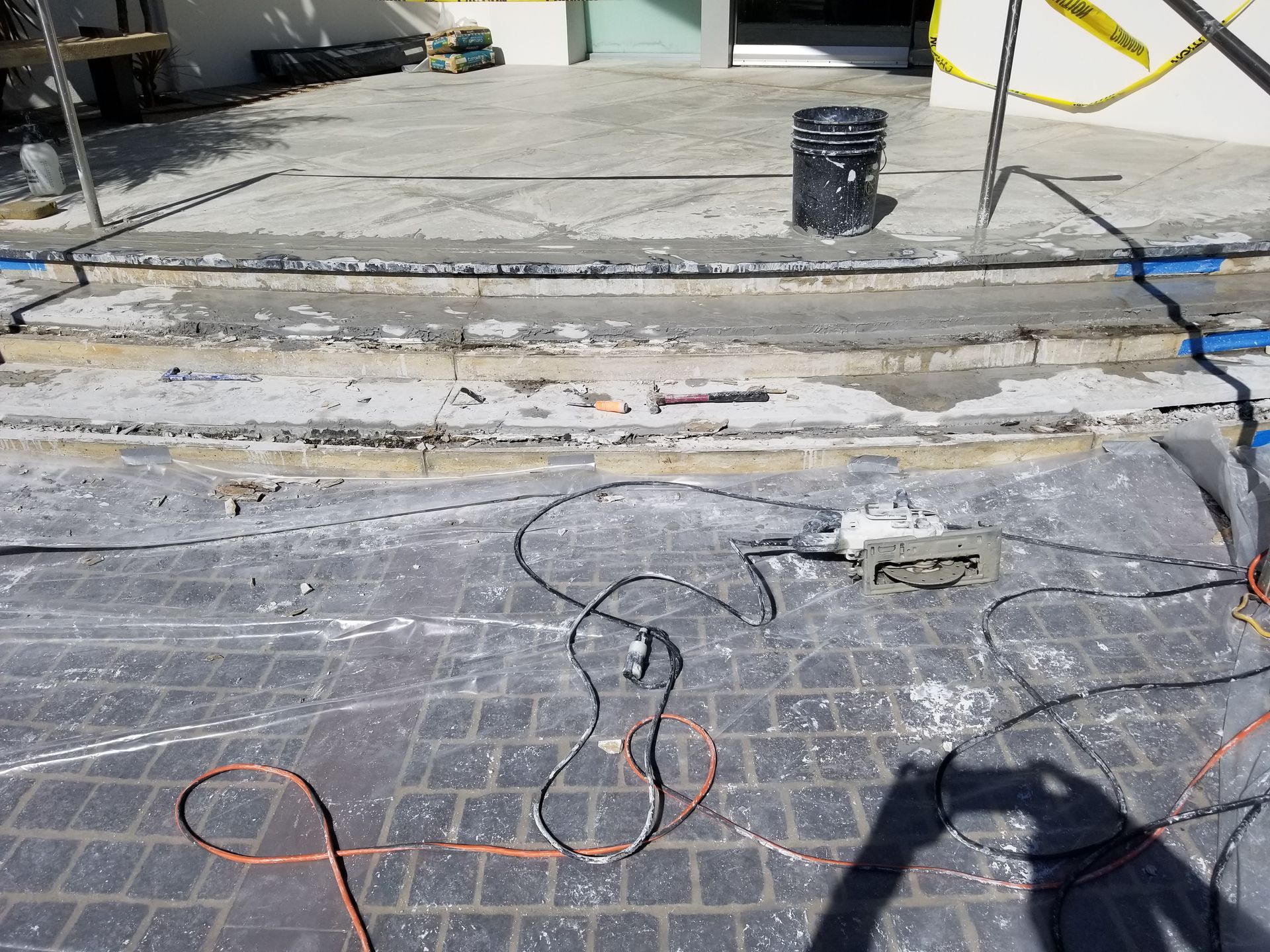 Construction site. Concrete steps being demolished. Power tools, wiring, and caution tape are present.