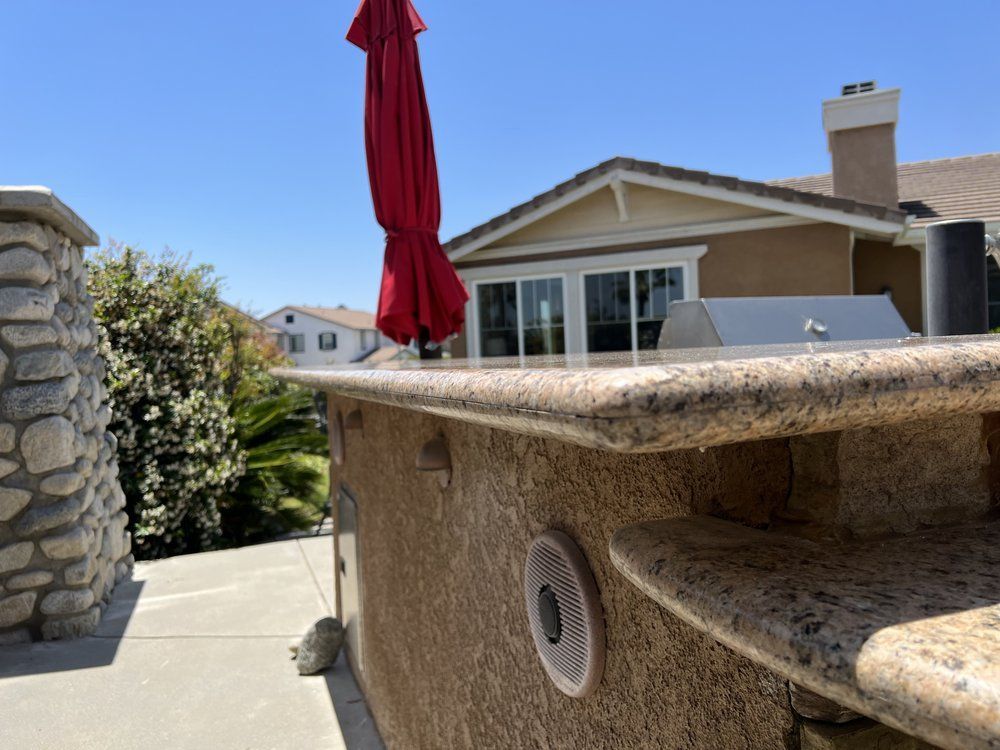 Outdoor kitchen with granite countertop, speaker, and red umbrella. House and sunny blue sky in background.