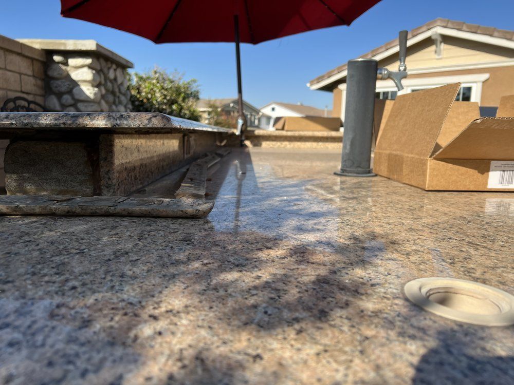 Granite countertop with a red umbrella, a built-in grill, and a suburban house in the background under a clear sky.