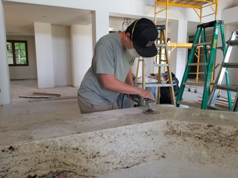 Man sanding countertop in a room under construction, wearing a hat and mask. Scaffolding and ladder in background.