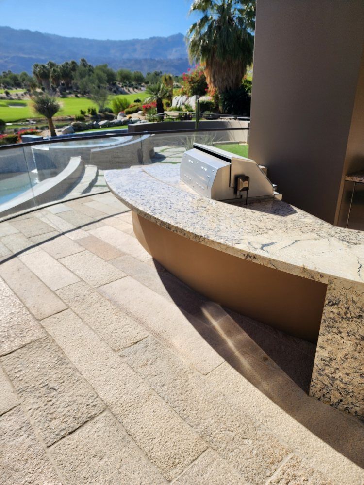 Outdoor kitchen with grill, light-colored granite countertop, beige base, stone patio, and mountain backdrop.