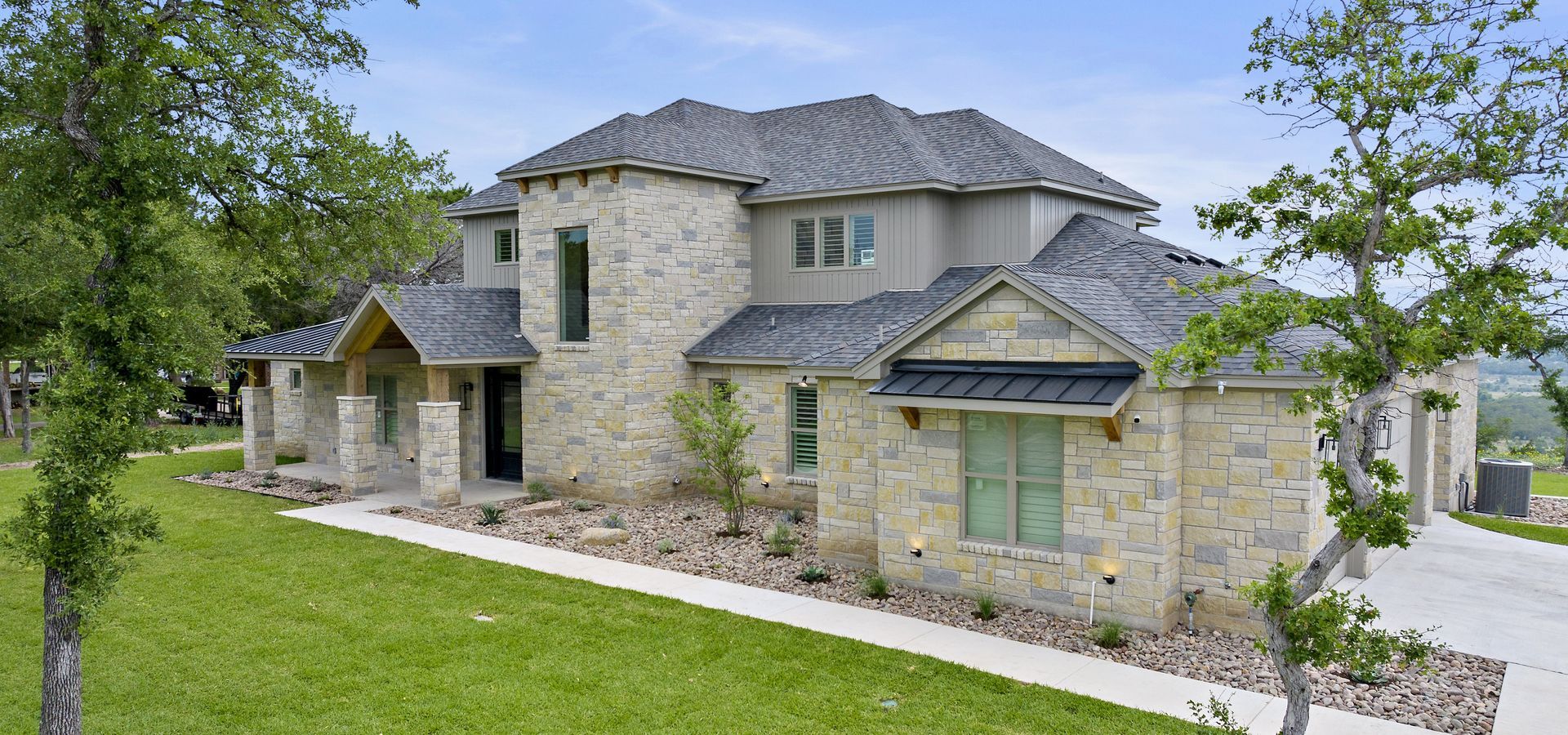 A two-story house with stone siding and a dark gray roof, surrounded by green lawns and mature trees under a blue sky.