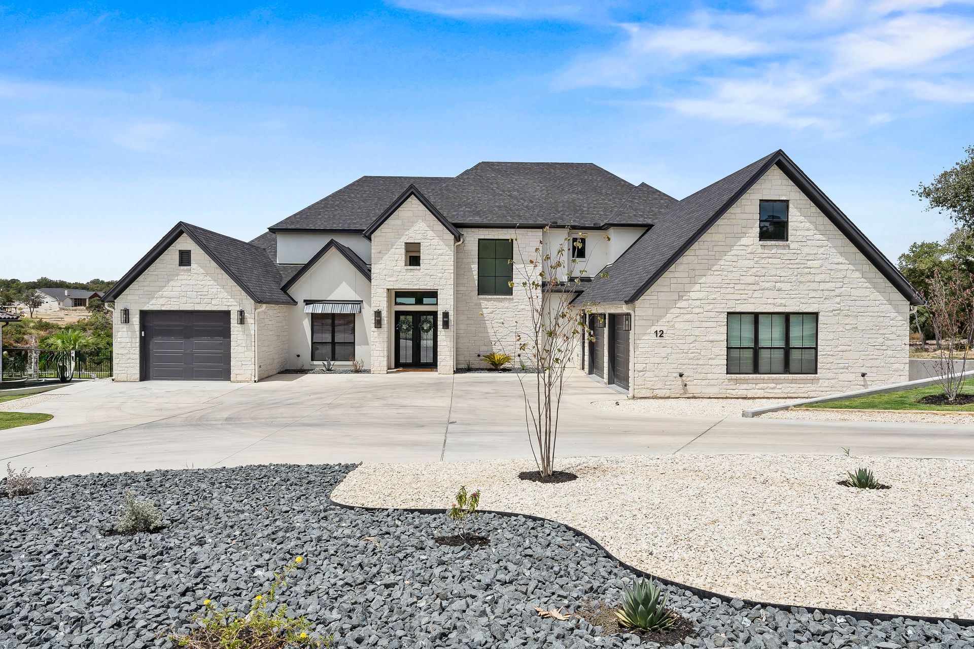 A large, light-stone house with a dark gray gabled roof, a wide concrete driveway, and landscaped rock beds.